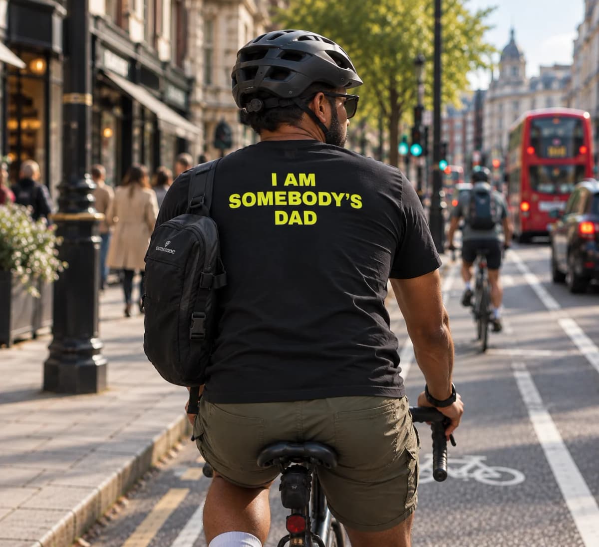 A man with a backpack cycling in a London bike lane, black t-shirt reading “I am somebody’s dad” in yellow